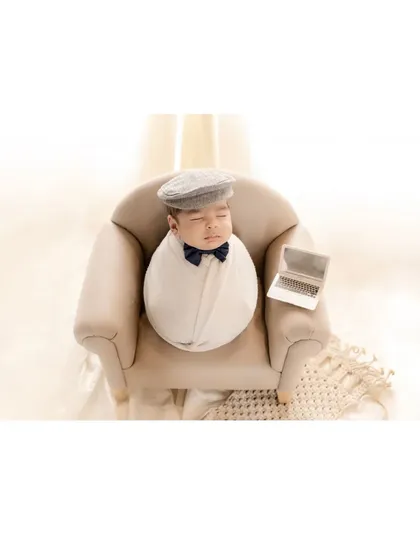 The little boss. A newborn, swaddled and wearing a dapper cap and bow tie, sleeps in a miniature armchair with a tiny laptop. This humorous and adorable themed shot shows the creative possibilities of a newborn session.