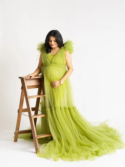 A full-length portrait of an expecting mother in a flowing lime green gown. She poses next to a small wooden ladder, adding a simple prop to this elegant studio shot.