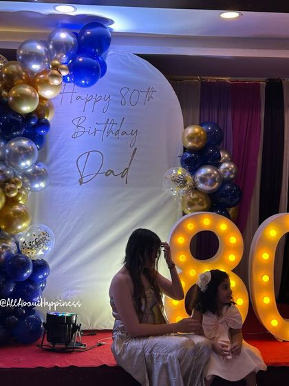 An elegant setup for an 80th birthday celebration, featuring a white arch backdrop with custom calligraphy, a blue, gold, and silver balloon garland, and large marquee numbers.