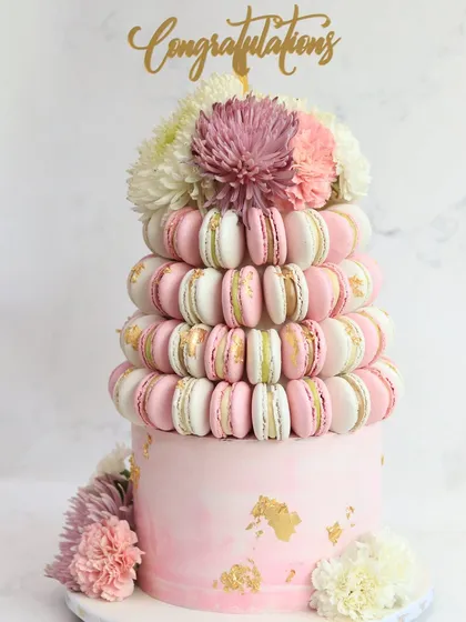 A stunning two-tiered celebration cake, featuring a base tier elegantly wrapped in a tower of handcrafted pink and white macarons. Topped with fresh flowers and a "Congratulations" sign, this design showcases our unique and bespoke cake artistry.