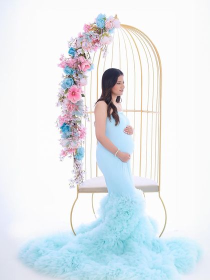 This whimsical portrait features a mother-to-be in a light blue gown, seated in a golden birdcage chair adorned with flowers. It's a perfect example of our creative, fairy-tale-inspired studio setups.