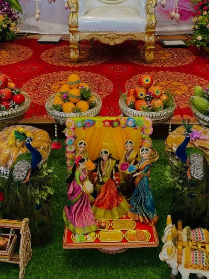 A close-up of the central part of the offering table. The arrangement features a traditional doll scene, flanked by peacock figures and surrounded by bowls of fresh fruits and sweets.