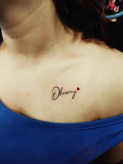 The name 'Dheeraj' with a tiny red heart on the collarbone. The placement and fine-line script make this a very personal and stylish tattoo.