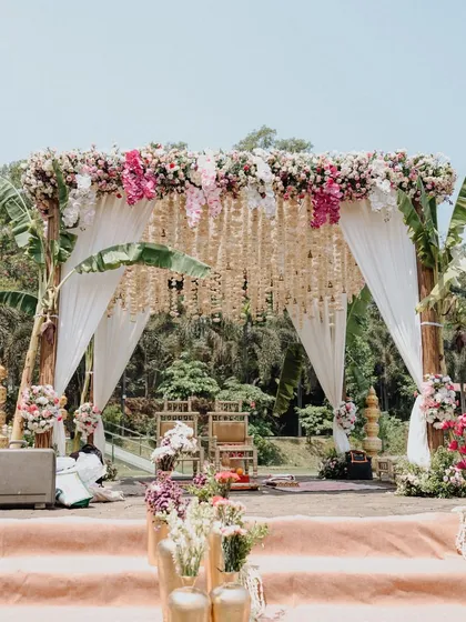 A closer look at a beautiful lakeside mandap. The structure combines rustic wooden poles with delicate drapes and a stunning overhead arrangement of pink and white flowers, complemented by traditional tuberose hangings.