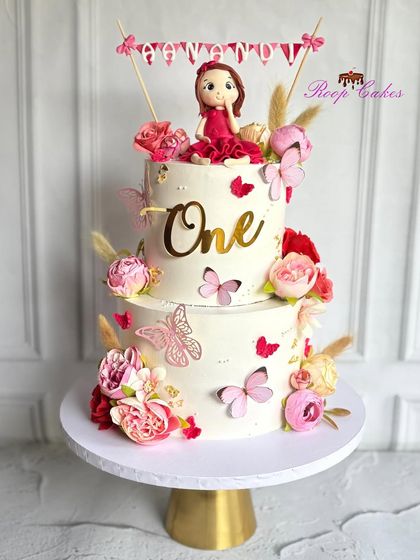 A beautiful two-tier cake celebrating "One." This whimsical design is decorated with pink and red flowers, butterflies, and a handcrafted doll topper, making it a perfect centerpiece for a first birthday.