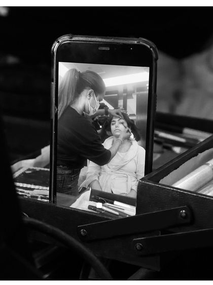 An artistic black and white photo taken over the shoulder of an artist, showing the client's makeup application reflected on a phone screen. It highlights our modern approach and attention to detail.