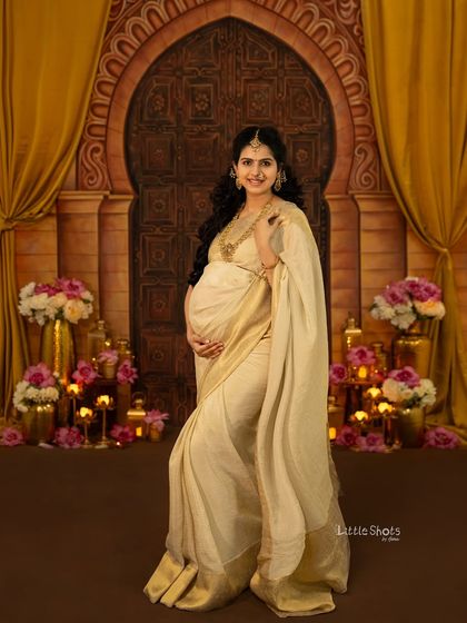 A grand and traditional portrait of an expecting mother in a cream and gold saree. She stands in a beautifully decorated room with an arched doorway, flowers, and candles, looking absolutely regal.