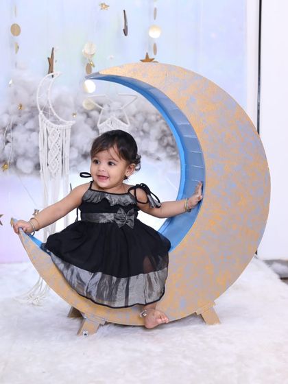 A smiling baby girl on the moon prop. This image highlights the "Celestial Dreams" setup, complete with hanging stars and a dreamcatcher for a bohemian touch.