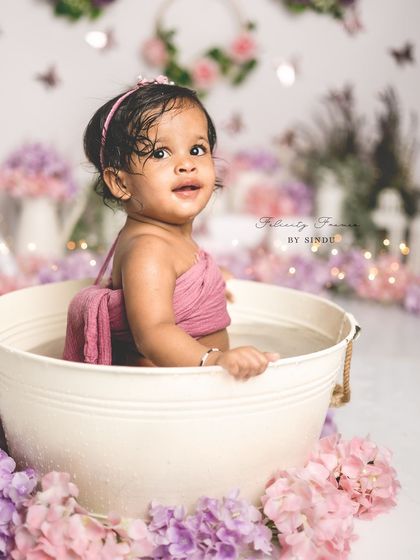 A happy baby girl in a tub, surrounded by purple and pink flowers. The splash session is the perfect way to clean up and have fun after a cake smash.