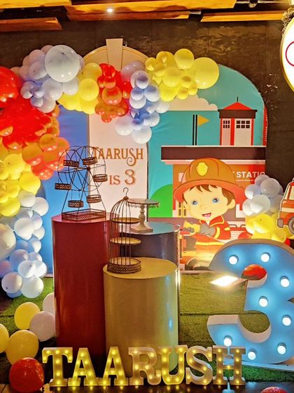 An exciting Fireman-themed third birthday party. The decor includes a fire station backdrop, firefighter character cutouts, and a large light-up number '3'.