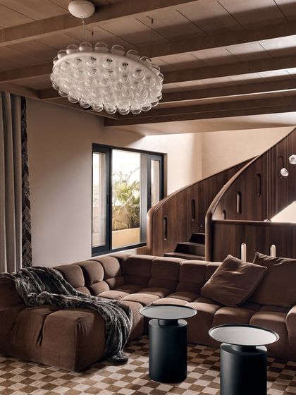 The main living area, featuring a comfortable, modular brown sofa and a stunning glass bubble chandelier. The space is anchored by a grand, curving wooden staircase, creating a sense of warmth and modern luxury.