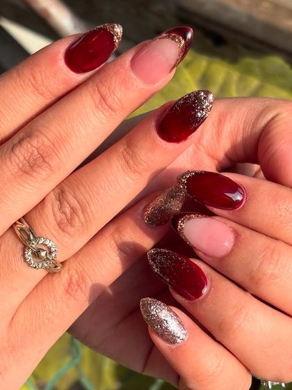 A dramatic and luxurious almond-shaped manicure, mixing deep maroon, sheer nude, and full-coverage gold and maroon glitter nails.