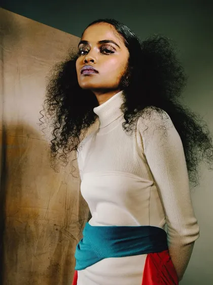 A portrait from the 'ANAINA' series against a neutral canvas backdrop. The styling is simple, allowing the model's voluminous hair and confident expression to be the main focus.