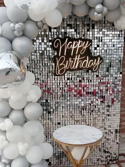 An outdoor birthday party with a silver sequin wall. The setup includes a white and silver balloon garland and a small table for the cake.