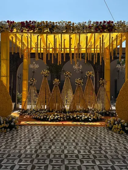 A grand mandap for an outdoor Haldi ceremony. The structure is draped in yellow fabric with hanging floral strings and chandeliers, complemented by large floral teardrop shapes.