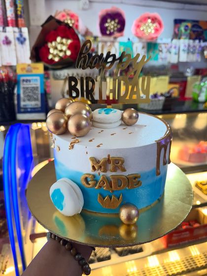 A custom cake for Mr. Gade, featuring a blue and white design with golden spheres and a crown accent. It's a stylish and personalized cake fit for a king.
