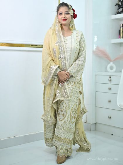 A full-length view of a model in a beautiful ivory sharara suit, perfect for a Nikkah ceremony or a pre-wedding event. The look is accessorized with a traditional passa and chandbali earrings.