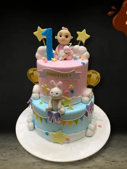 A delightful two-tier cake celebrating a first birthday. The pink and blue pastel design is decorated with stars, clouds, and adorable fondant bunnies, creating a dreamy and sweet theme for the special day.