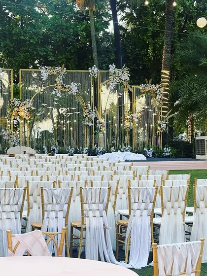An elegant daytime reception setup with a mirrored gold backdrop. The design reflects the surrounding palm trees and sky, creating a unique and glamorous effect for an outdoor event.