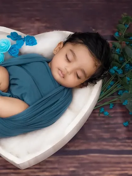 A baby wrapped in blue rests in a white heart-shaped bowl, complemented by blue flowers and a rustic wood background.