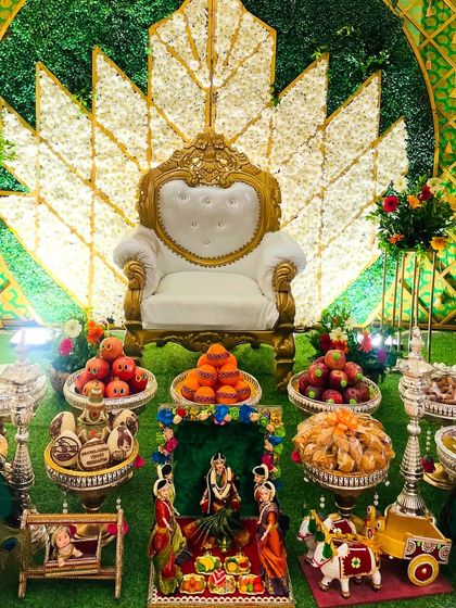 A beautiful lotus-themed backdrop made with white and green florals for a baby shower. The setup is completed with peacock props, a traditional doll scene, and plates filled with colorful fruits and sweets.