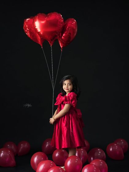 A striking portrait of a girl in a beautiful red dress holding red heart-shaped balloons against a solid black background.