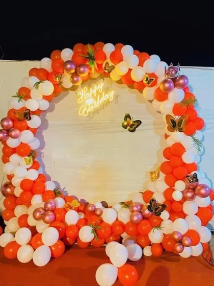 A bright and cheerful birthday backdrop in orange and white. The circular balloon arch is decorated with butterfly accents and a 'Happy Birthday' neon sign.