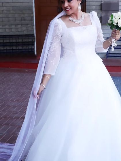 A radiant bride on her wedding day, holding her bouquet. Her custom A-line gown with sheer embroidered sleeves and a long veil creates a look of pure elegance and happiness.