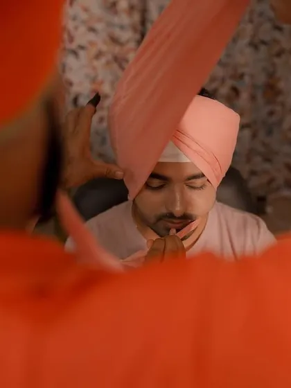 An artistic view of the groom during the turban tying ceremony, framed by the vibrant orange of a family member's attire.