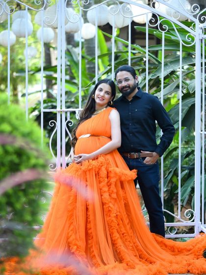 A stylish couple portrait in front of an ornate white gate. The mom-to-be's orange gown is a beautiful centerpiece.