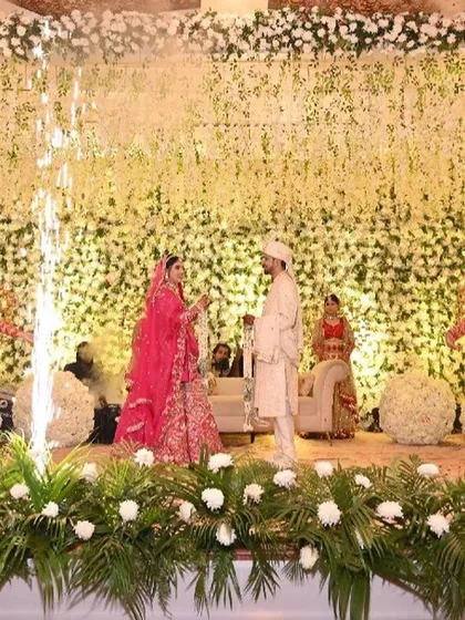 A mesmerizing Jaimala ceremony with the couple on a stage decorated with lush greenery and white flowers. Multiple pyrotechnic fountains and smoke jets create a celebratory and grand atmosphere.