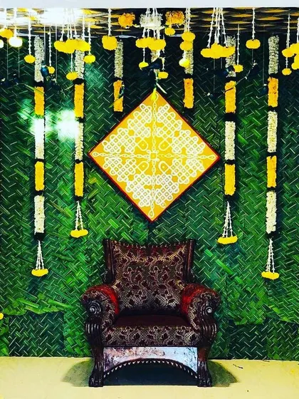 A traditional backdrop for a ceremony, featuring a woven mat background, hanging flower garlands, and a central kolam or rangoli art piece.
