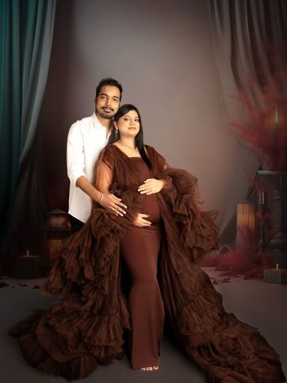 A warm and intimate couple's portrait. The expecting mother is wrapped in a luxurious brown ruffled gown while her partner embraces her from behind in a softly lit studio setting.