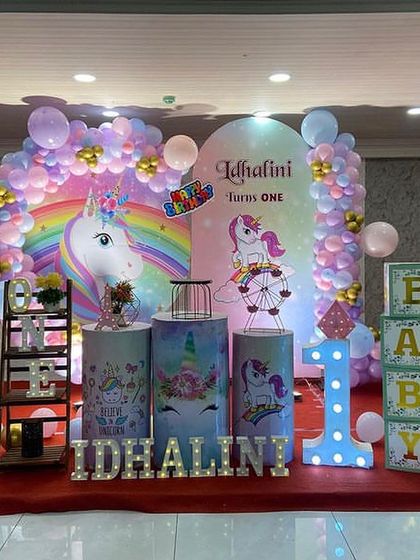 A complete unicorn stage setup for a first birthday, with rainbow backdrops, light-up letters and number, and themed pedestals.
