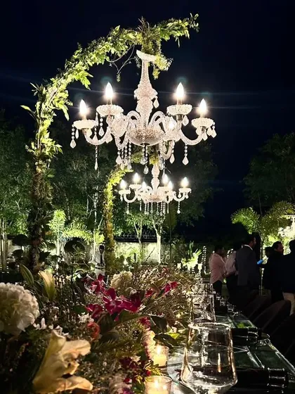 An enchanting outdoor sit-down dinner under the stars. We used greenery-wrapped chandeliers to create a canopy of light and elegance over a long, mirrored table, crafting an intimate and utterly romantic dining experience.