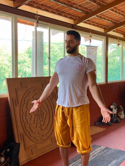 A student stands in a moment of quiet contemplation, feeling the energy of the shala. The wooden mandala behind him is a tool for meditation, representing the journey to the center of the self.