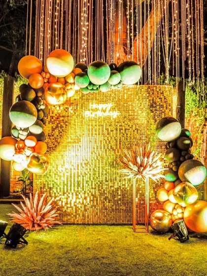 A dazzling photo booth perfect for a sangeet or cocktail party. This outdoor setup includes a shimmering gold sequin wall, a stylish balloon garland in metallic tones, and unique floor lamps, all under a canopy of hanging fairy lights.