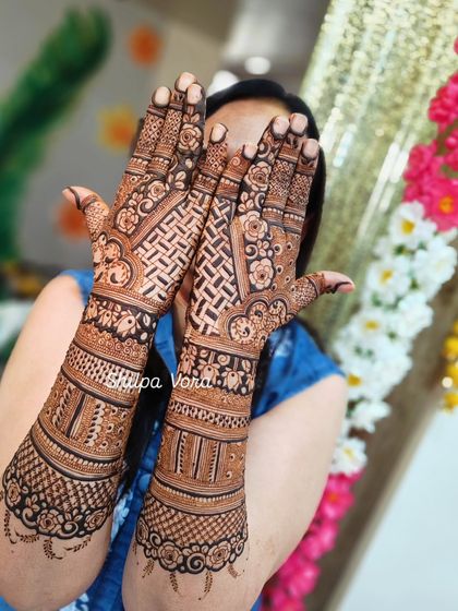 A happy bride showing off her elaborate full-arm mehndi against a festive floral backdrop.