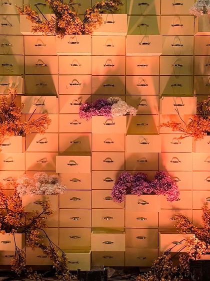 A creative and unique photo backdrop for a Haldi ceremony. This wall is constructed from stacked wooden boxes, with clusters of orange and purple flowers adding pops of color.