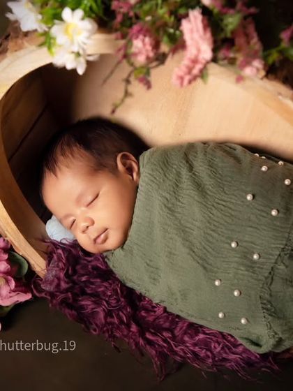 A detailed close-up shot of the baby in the bark bowl, showing the delicate pearl details on the green swaddle and the soft floral accents.