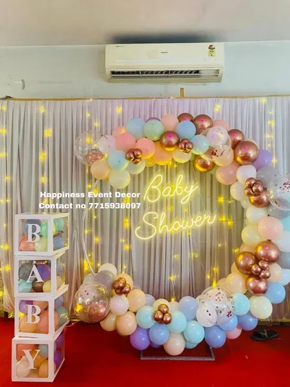 This modern baby shower setup is very popular. It features a full ring backdrop covered in a garland of pastel and metallic balloons, a 'Baby Shower' neon sign, and transparent 'BABY' boxes filled with matching balloons.