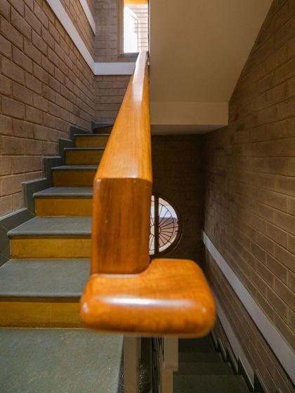 Detail of the staircase handrail, crafted from polished wood. The walls along the staircase are made of exposed earth blocks, and a circular window brings in light, highlighting the natural material palette.