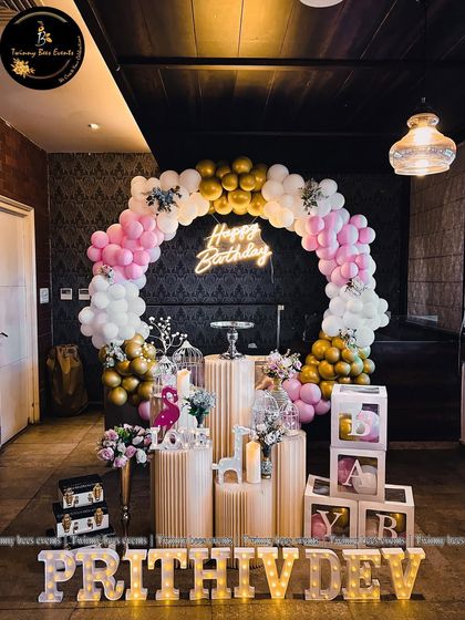 A full view of Prithivdev's first birthday setup. It features a circular balloon arch in pink, white, and gold, fluted pedestals, "BABY" blocks, and his name in large marquee letters.