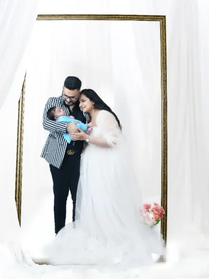 A creative family portrait where the new parents are holding their baby, framed by a large mirror. The white, dreamy setup creates an artistic and memorable first family photo.