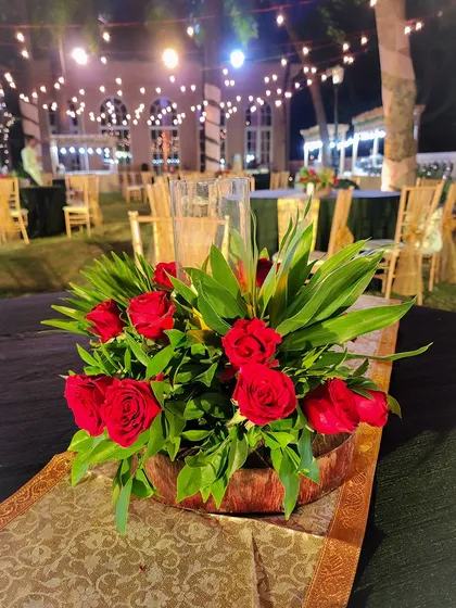 A simple yet classic centerpiece for an outdoor evening event. A low wooden bowl holds an arrangement of lush red roses and tropical green leaves, set on a patterned table runner.