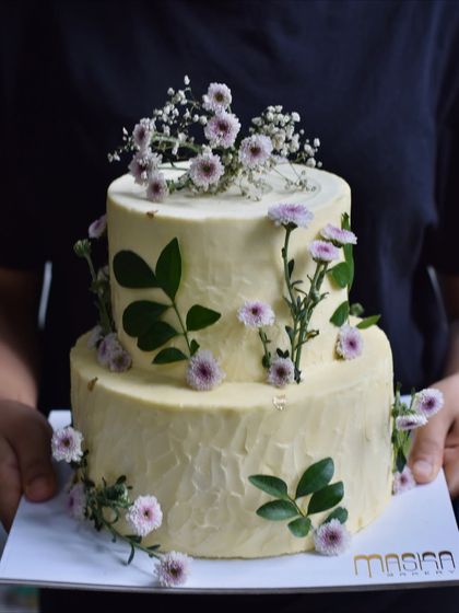 A beautiful two-tiered cake with a rustic buttercream texture, decorated with delicate purple asters and fresh green leaves. This design is perfect for an outdoor or bohemian-themed wedding or engagement party.