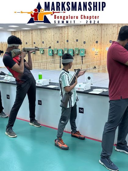 A dynamic action shot from the Open Sight Rifle competition, capturing the moment a shot is fired. The branding for the Marksmanship Summit is visible in the foreground.