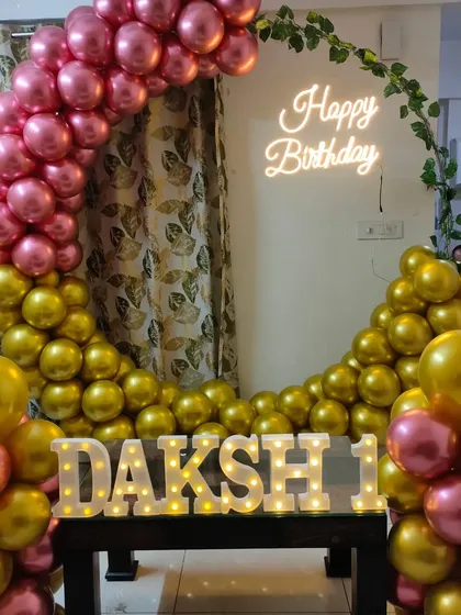 A metallic pink and gold balloon arch with a neon sign, set up for a first birthday at home.