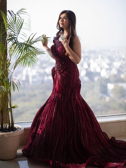 A client posing by a window in the red sculpted gown. The natural light highlights the intricate, wave-like texture of the fabric and its rich, deep color.