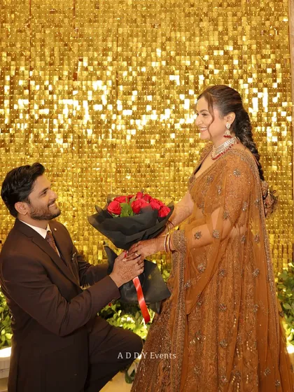 A romantic proposal moment during the engagement ceremony. The groom gets down on one knee against a glittering gold sequin backdrop, creating a picture-perfect memory.